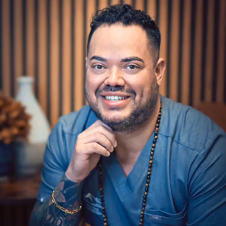 Smiling man with tattoos wearing blue medical scrubs and a beaded necklace in front of a wooden slat background.