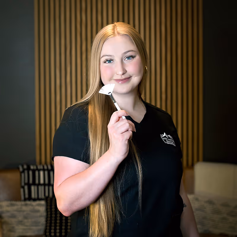 Smiling woman with long blonde hair holding a white fan makeup brush in a cosmetic setting.
