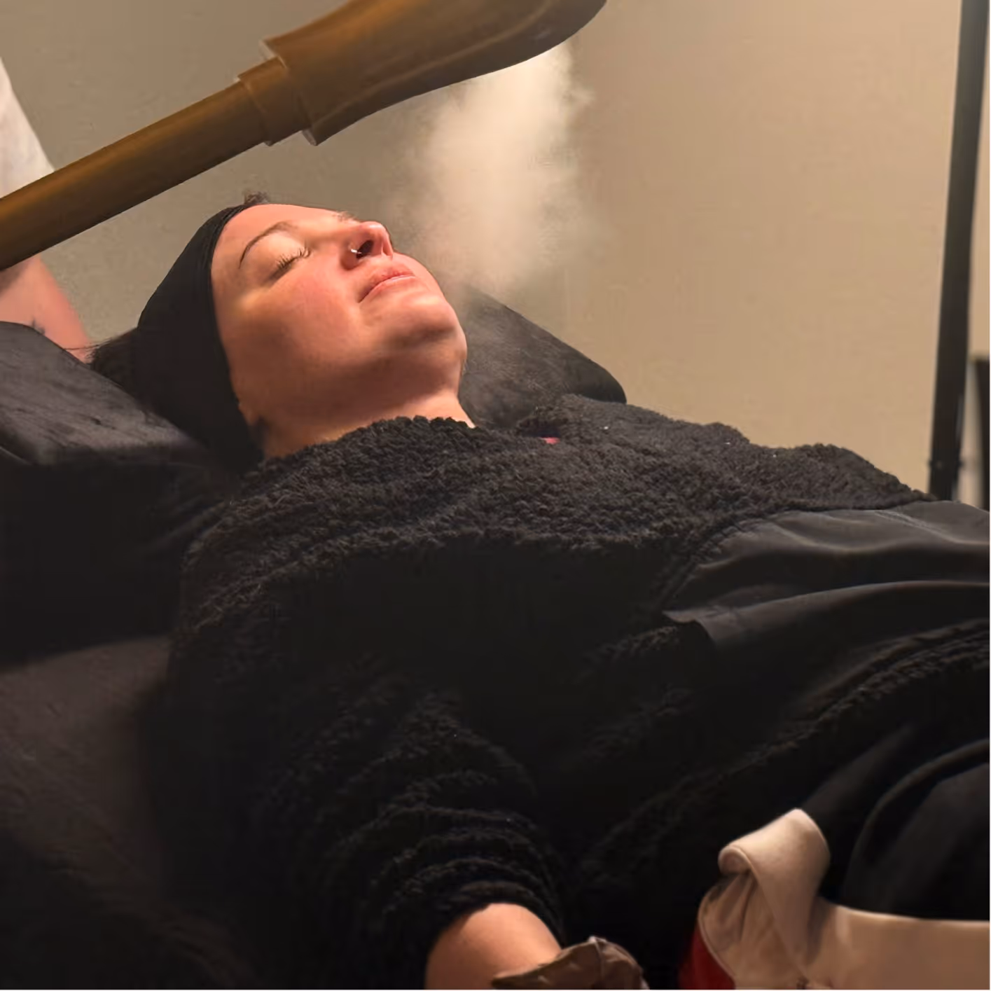 Woman with eyes closed receiving facial steam treatment while reclining on a spa bed.