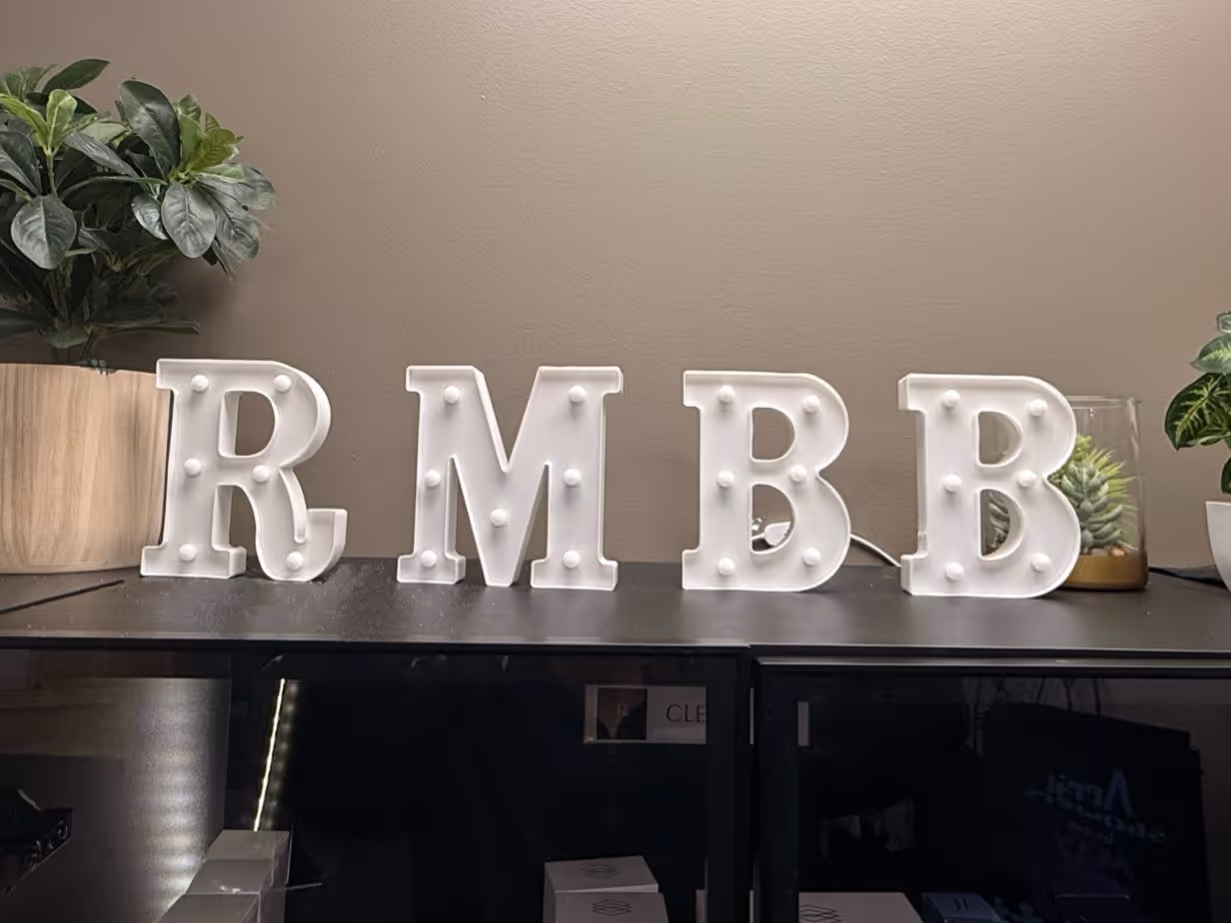 White marquee letters R M B B with small round bulbs, placed on a black surface with potted plants on either side.