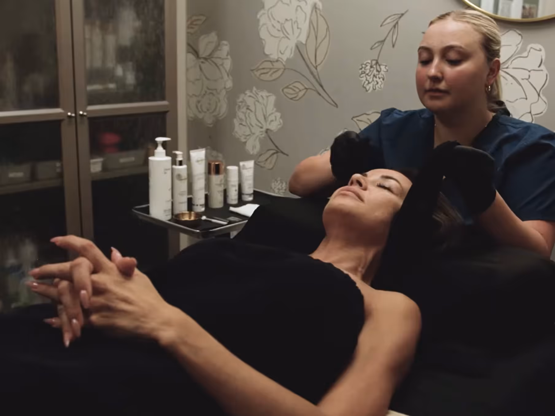 Woman receiving a facial treatment from an esthetician wearing black gloves in a spa setting.