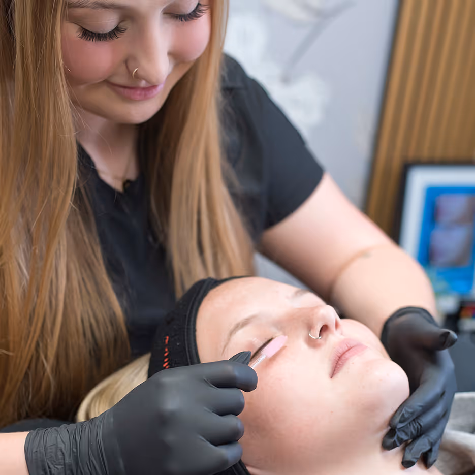 A cosmetologist wearing black gloves applies a beauty treatment to a woman's closed eyelid while the woman reclines with a black headband.
