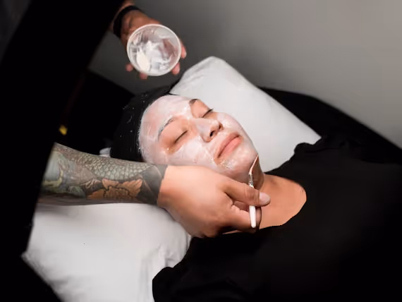 Person with eyes closed receiving a facial treatment with a white cream mask being applied with a brush by a tattooed arm.