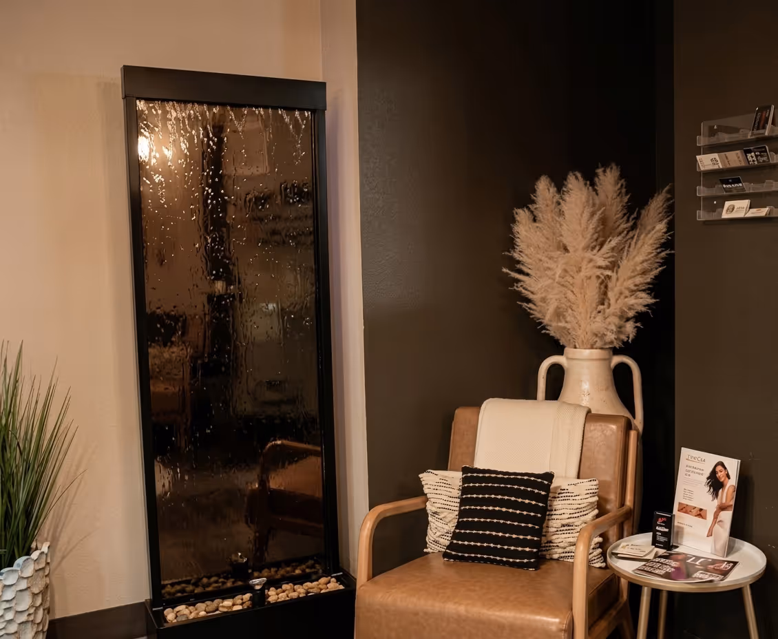 Cozy spa waiting area with a brown leather armchair adorned with two pillows and a blanket, a tall beige vase with pampas grass, a small round table with magazines, and a black water feature wall.