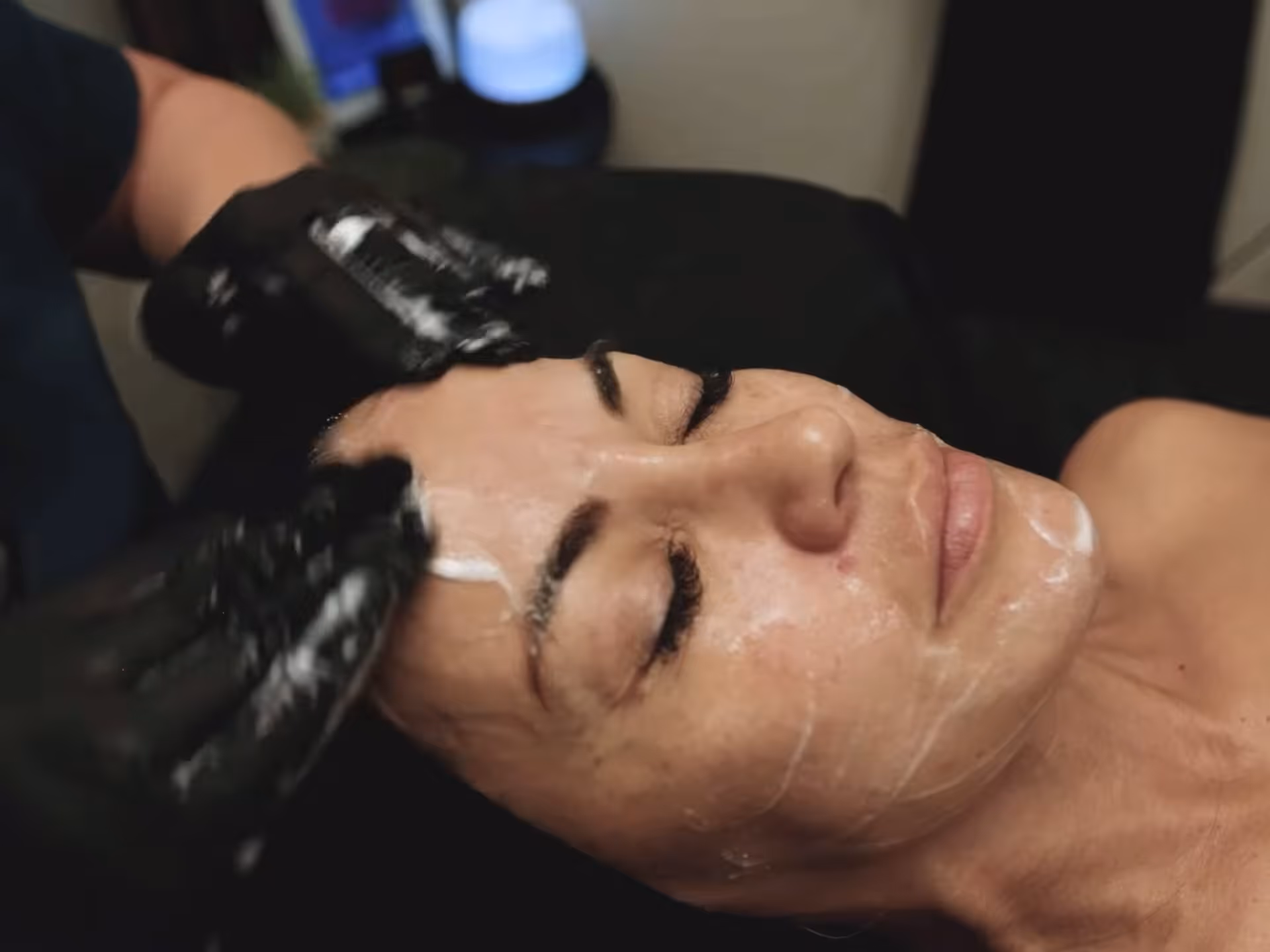 Woman receiving facial treatment with creamy cleanser applied on her face and a gloved esthetician massaging her forehead.