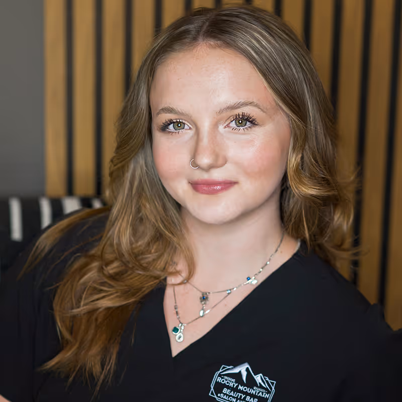 Smiling young woman with light brown wavy hair wearing a black shirt with Rocky Mountain Beauty Bar Salon & Spa logo.