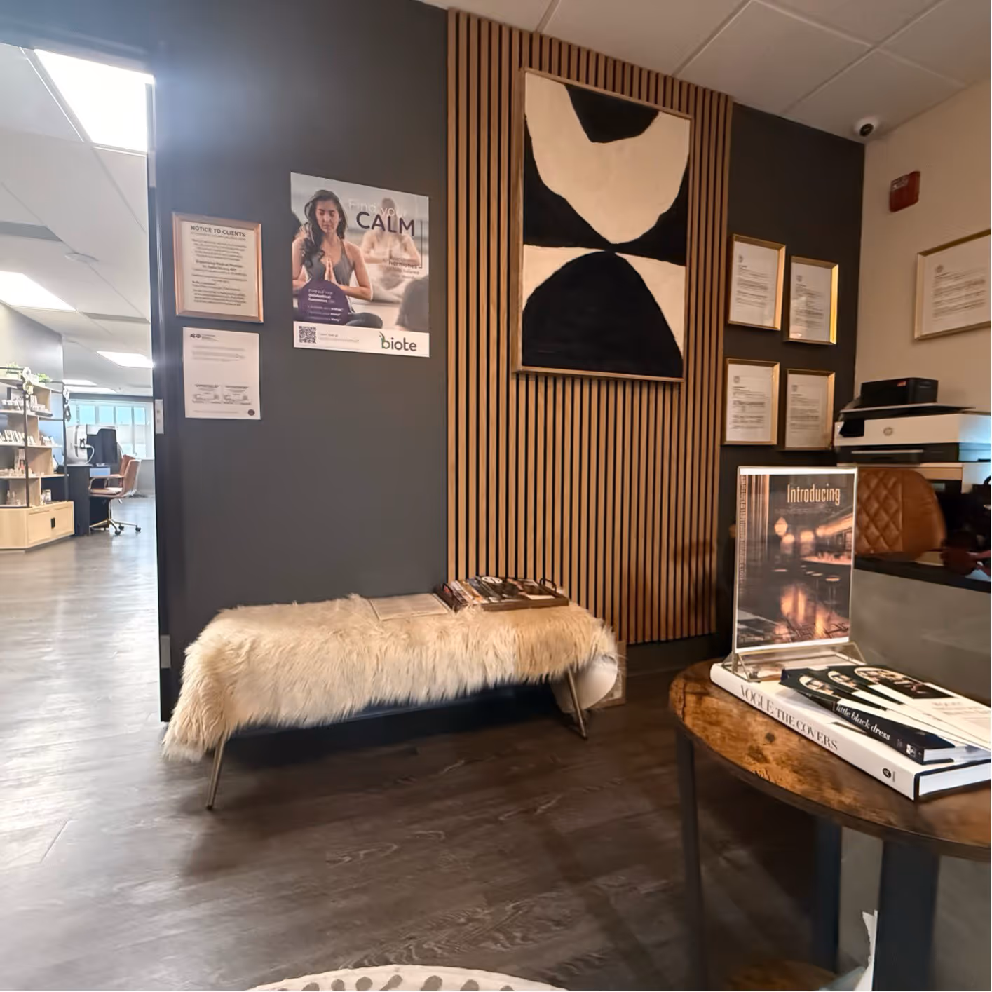 Modern spa reception area with a furry bench, abstract black and white wall art, framed certificates, and a round wooden table with books and a promotional display.