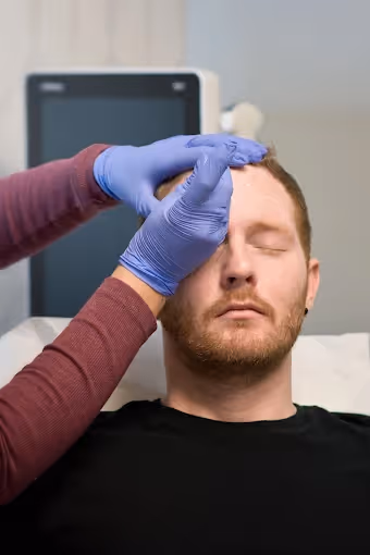 Man receiving forehead injection treatment with gloved hands in a clinical setting.
