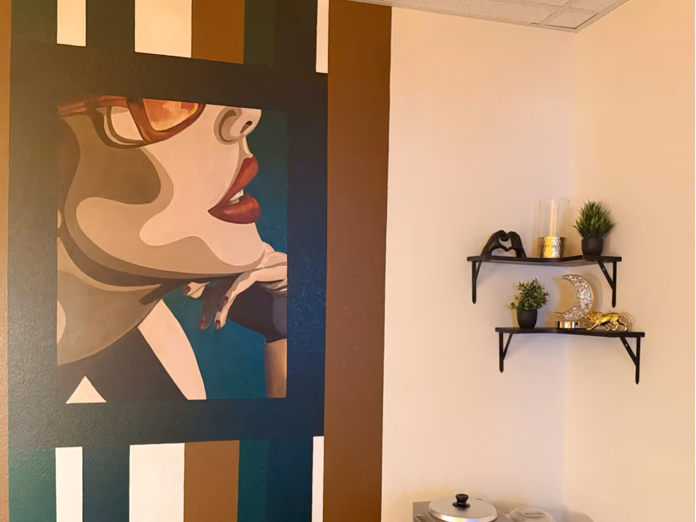 Wall mural of a woman wearing red lipstick and glasses resting her chin on her hand with two black shelves displaying plants and decorative items on a beige wall.