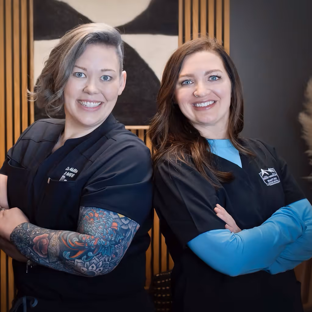 Two smiling women in black medical scrubs standing back to back with crossed arms, one with a colorful tattoo sleeve.