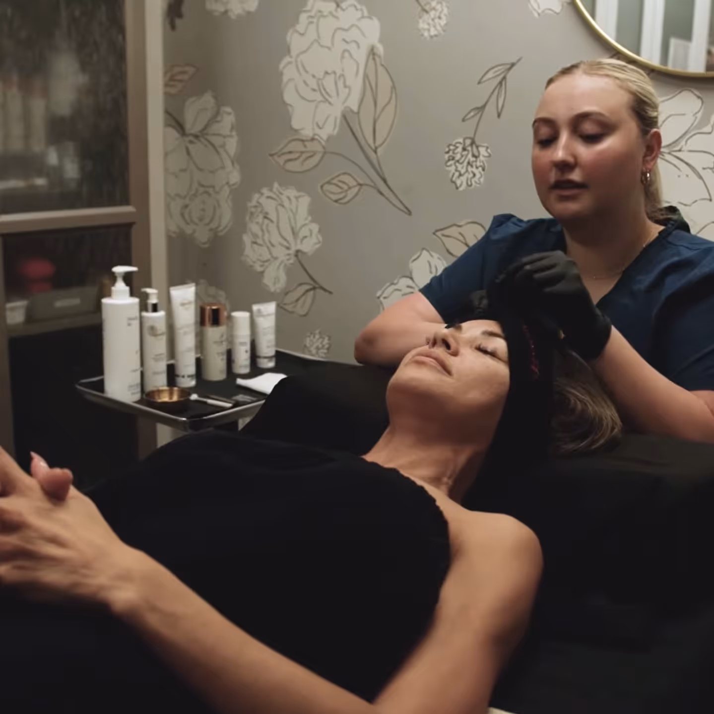 Woman receiving a facial treatment from a beauty professional wearing gloves in a spa room with skincare products on a nearby table.