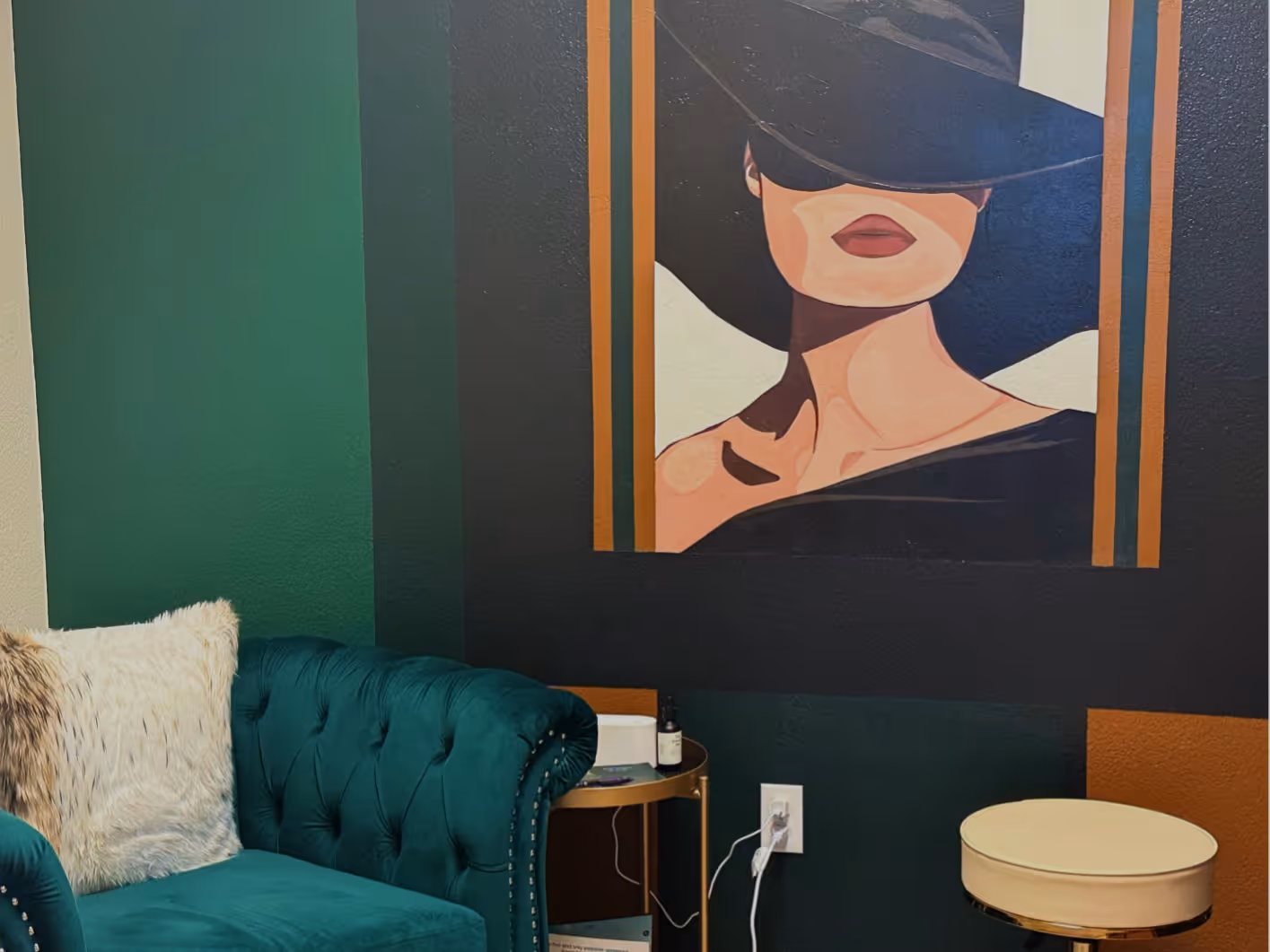 Cozy green velvet tufted armchair next to a round beige stool and a side table, with a large framed artwork of a woman wearing a black hat covering her eyes on the dark green wall.