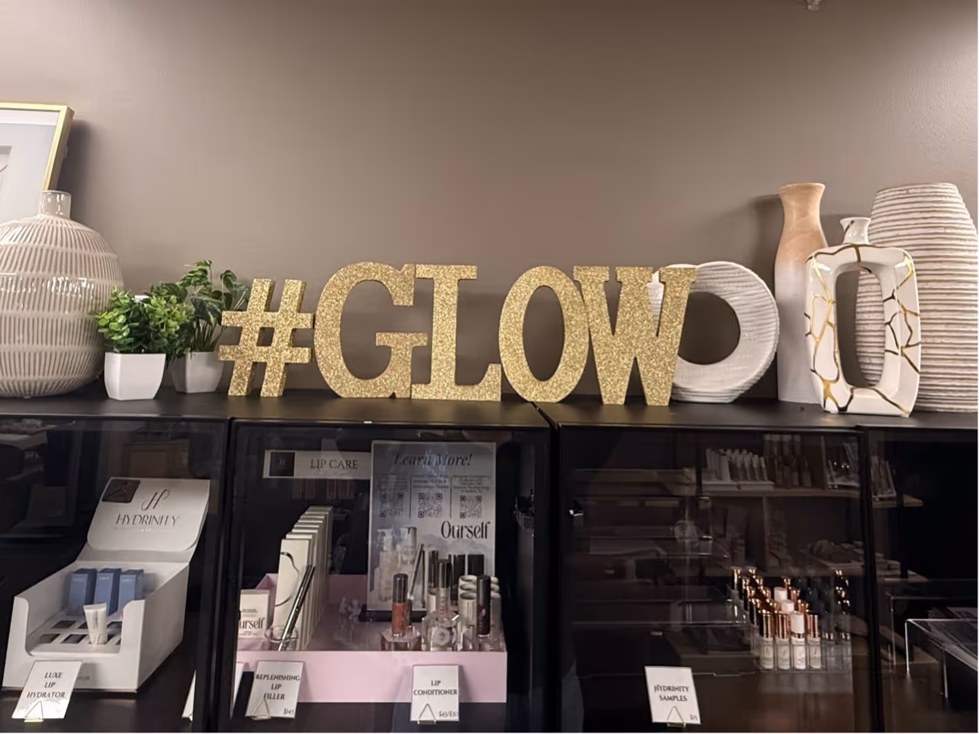 Shelf with gold glitter letters spelling #GLOW surrounded by decorative vases and small potted plants, with various lip care products displayed below.