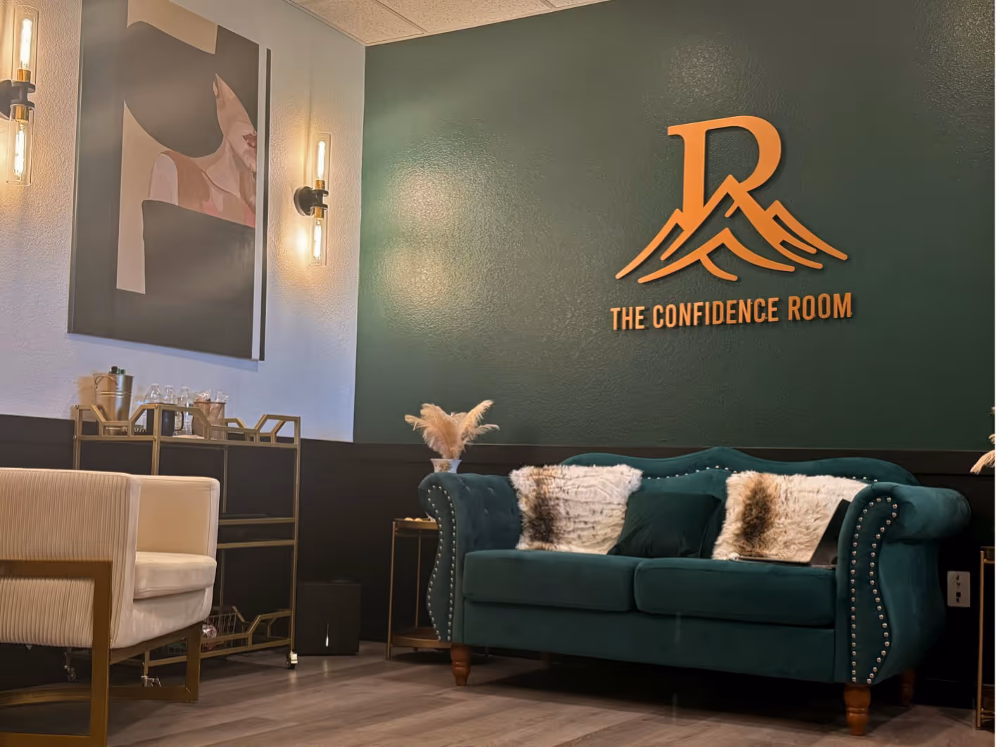 Reception area with green velvet sofa, beige armchair, wall art, and The Confidence Room logo on dark green wall.