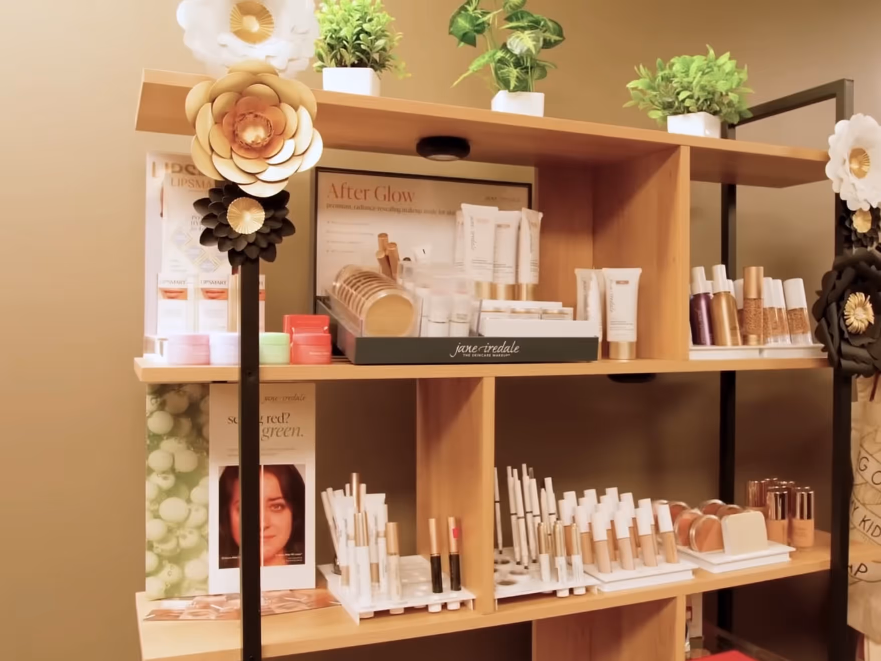 Wooden shelves displaying an assortment of skincare and makeup products, decorated with potted green plants and large decorative flowers.