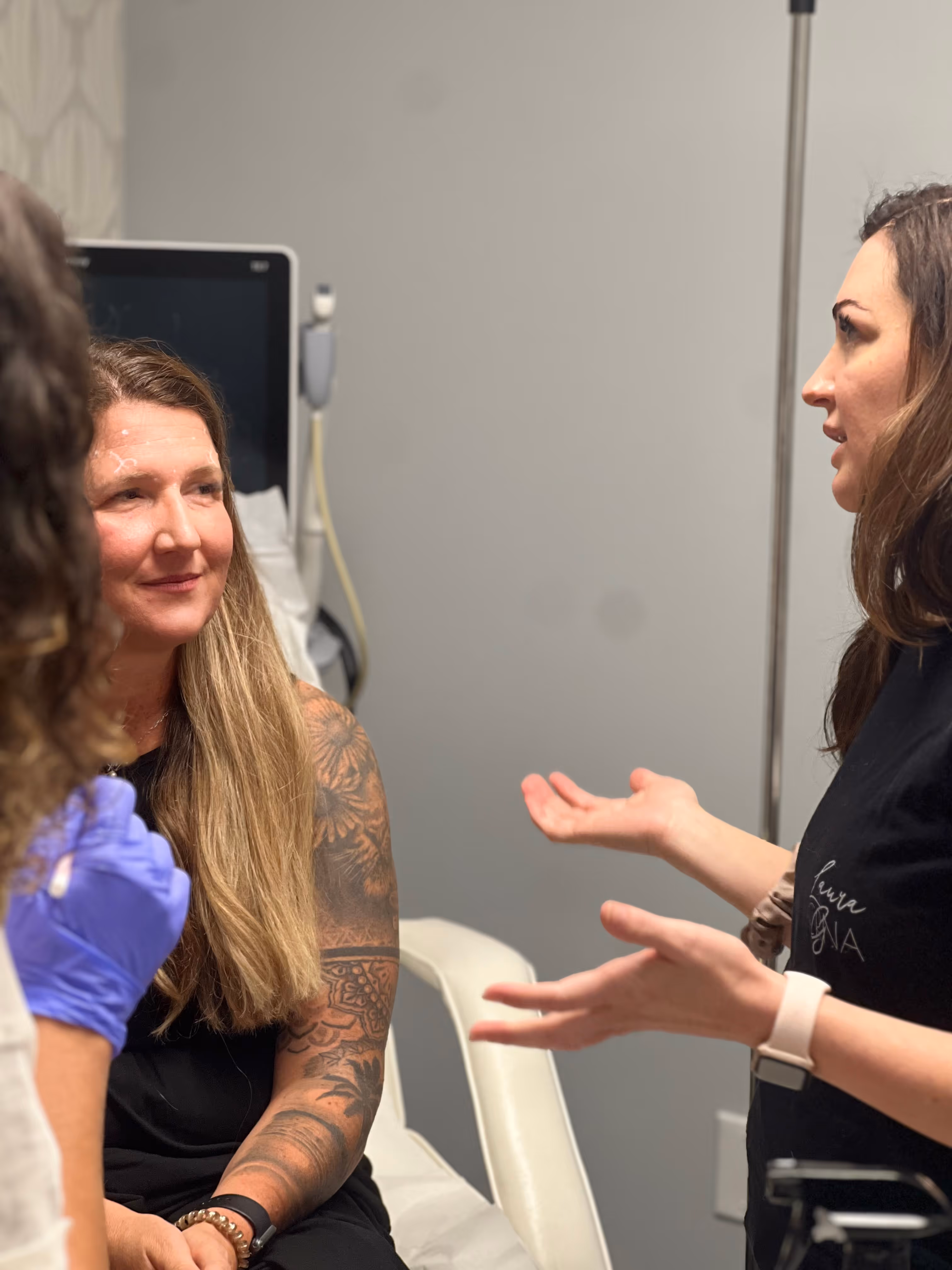 A medical professional explains forehead treatment markings to a tattooed woman sitting in a clinic chair.