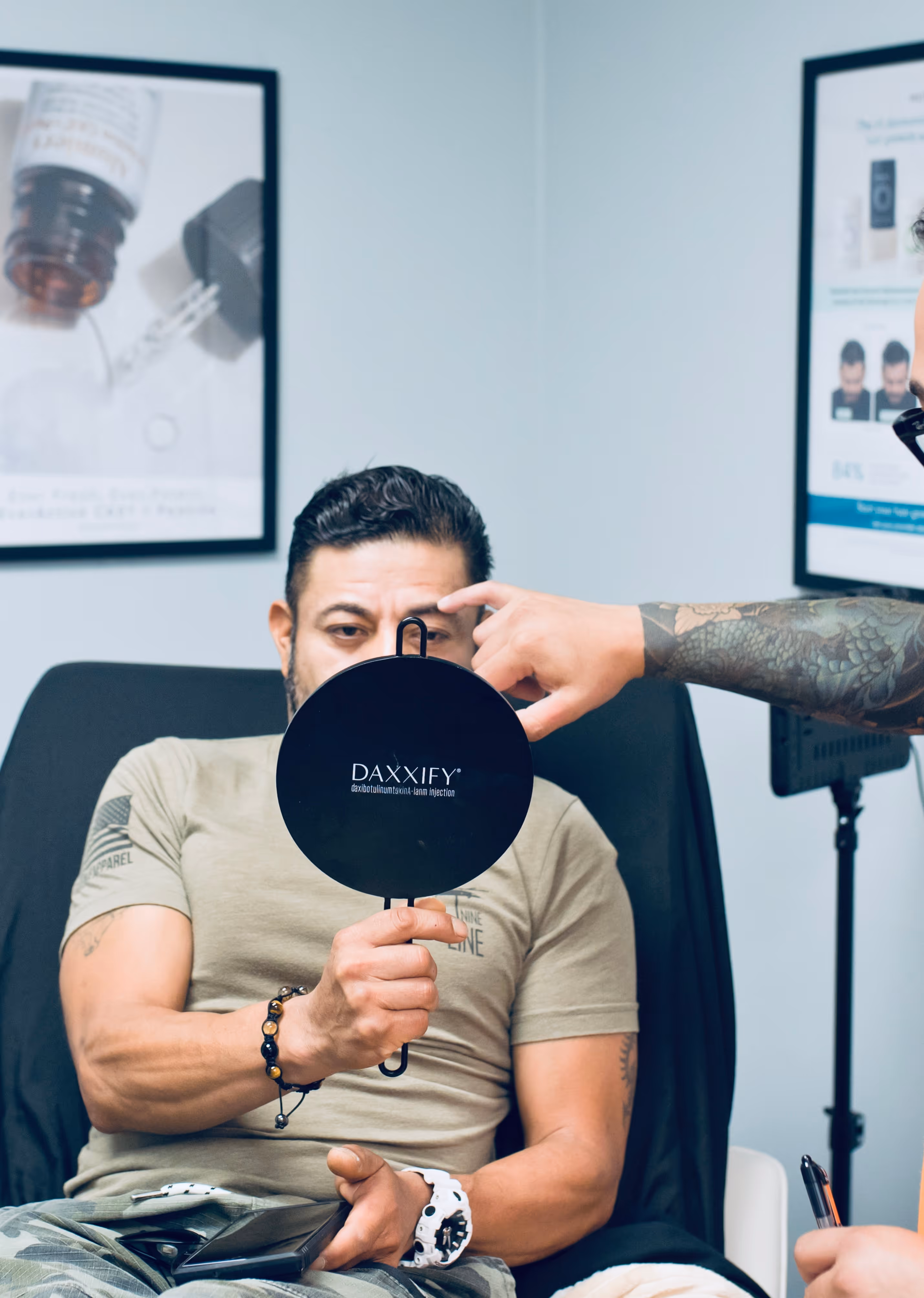 Man holding a black round mirror with 'DAXXIFY' text, while a person with a tattooed arm points at his forehead during consultation.