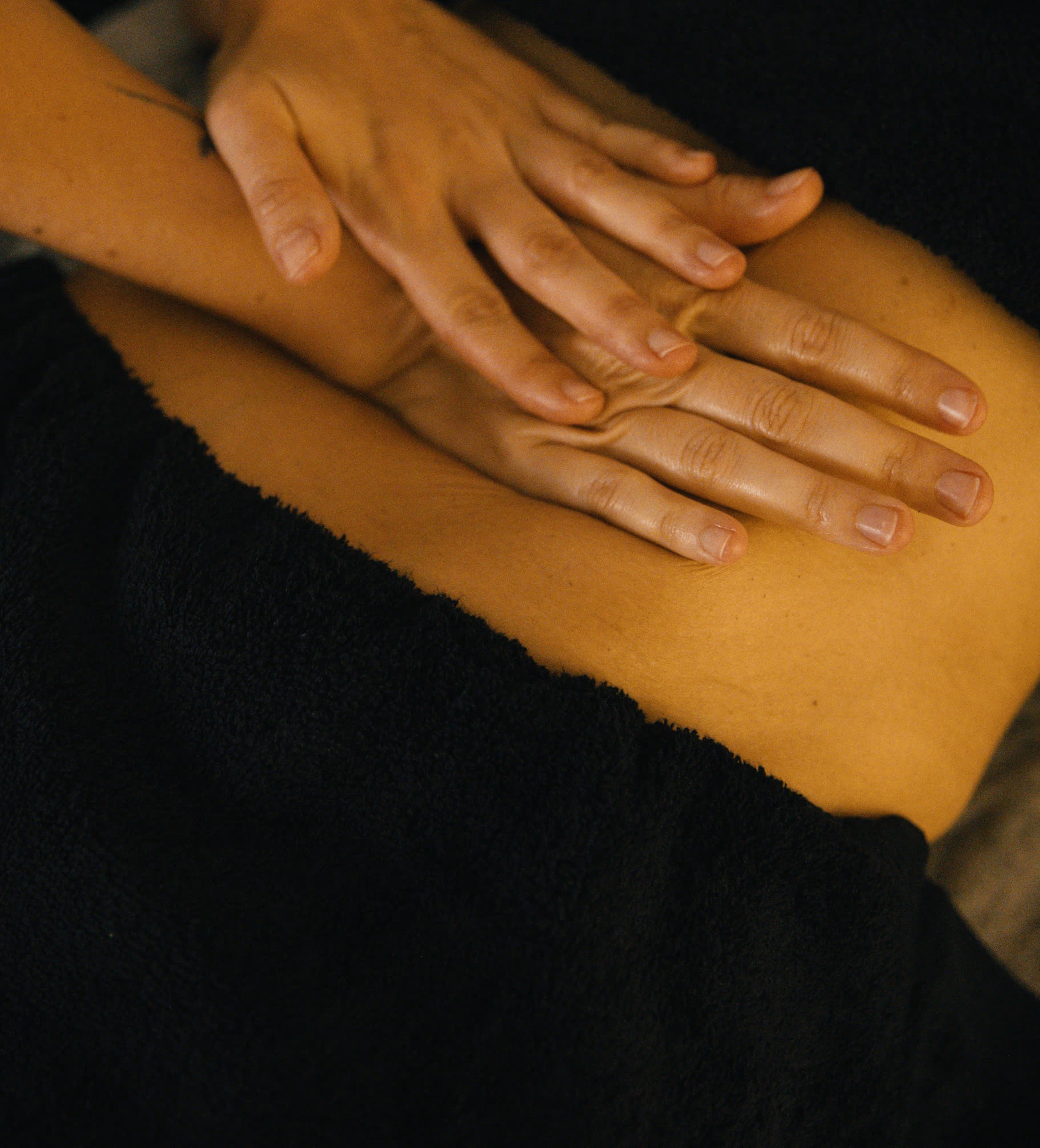 Close-up of two hands resting gently on a person's bare lower back over a black towel, suggesting a massage or wellness treatment.