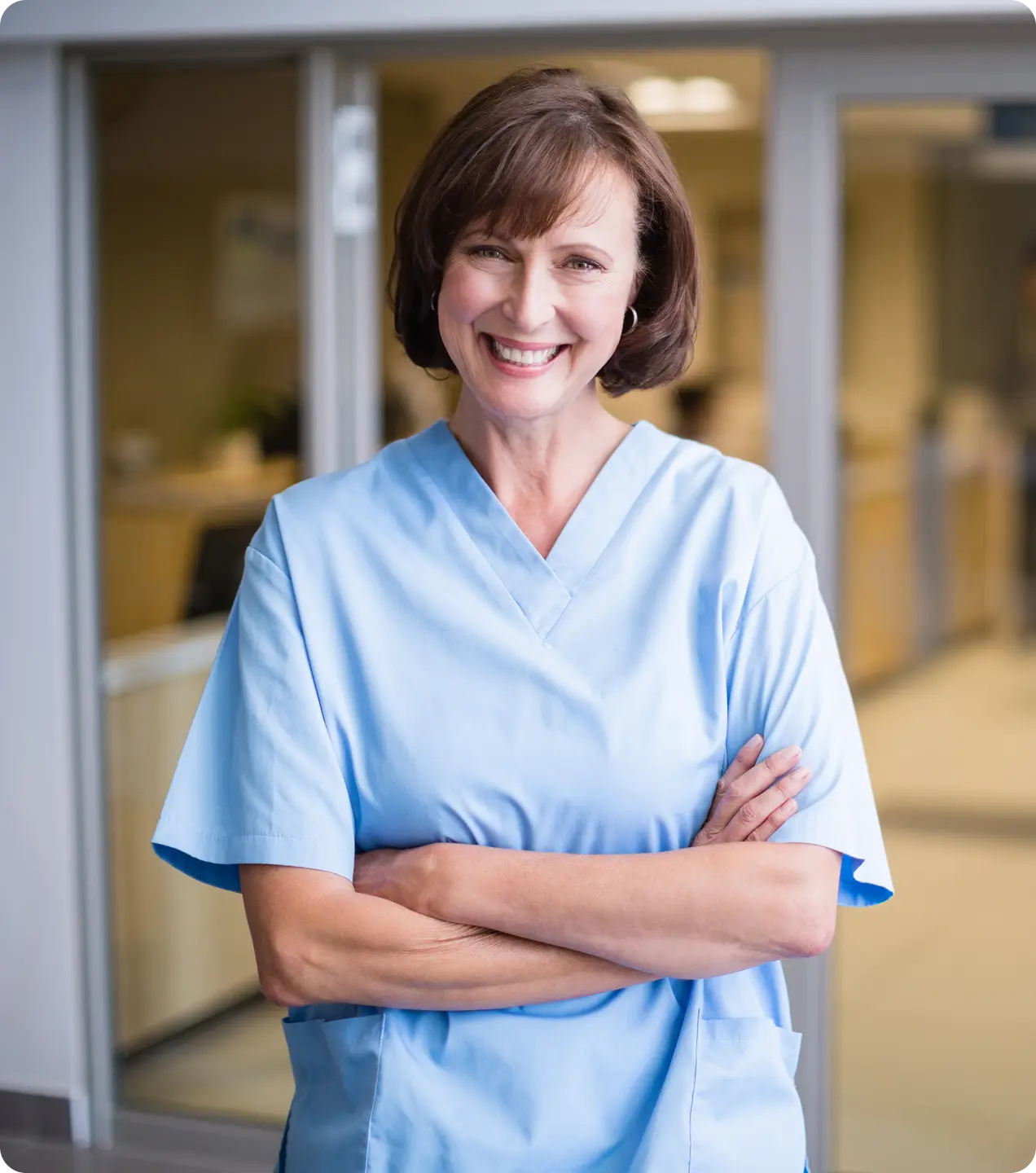 Smiling female nurse in light blue scrubs with arms crossed in a hospital corridor.