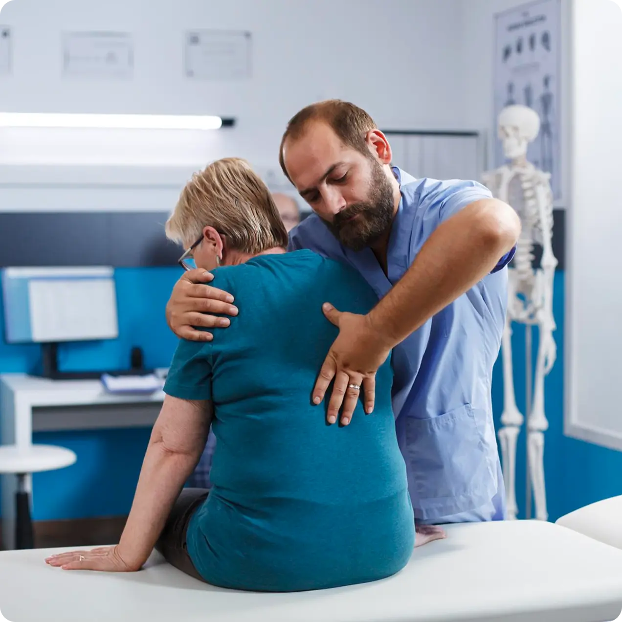 Male doctor in scrubs examining an elderly woman's back in a medical office with a skeleton model nearby.