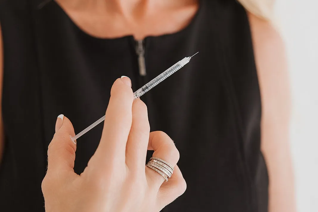Hand holding a small syringe with a needle, close to a person wearing a black top.