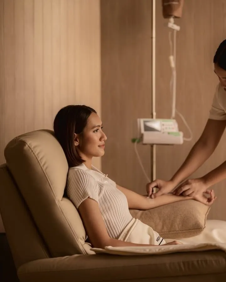 A woman receives an intravenous infusion while sitting in a beige recliner chair.