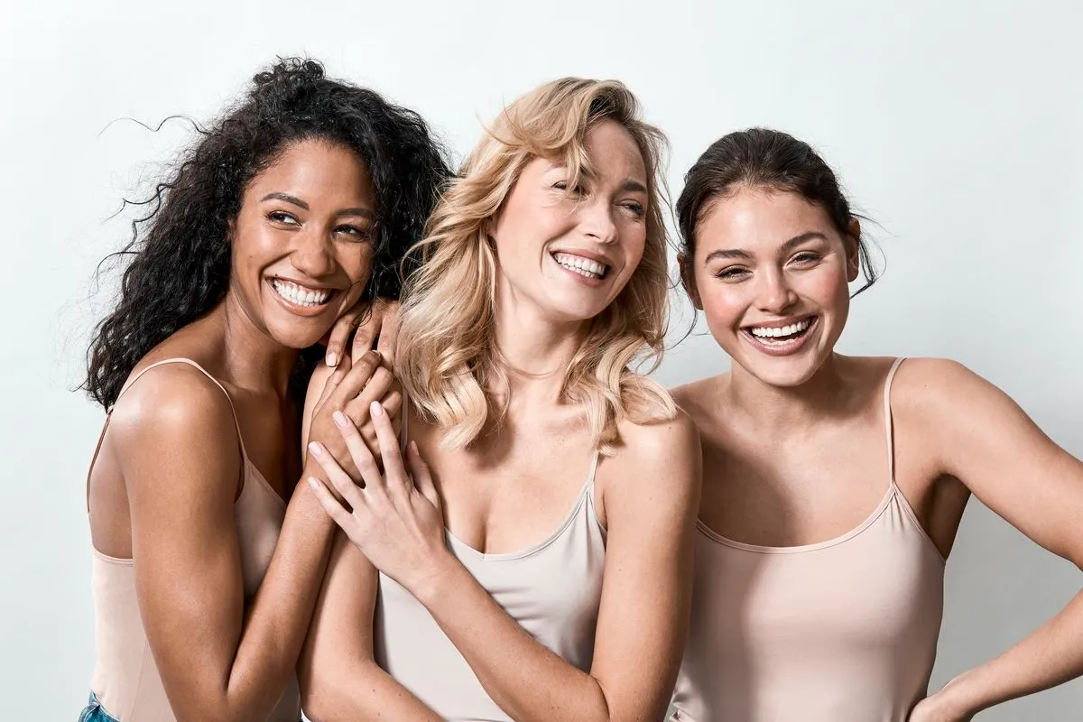 Three smiling diverse women in beige tank tops posing closely against a plain light background.
