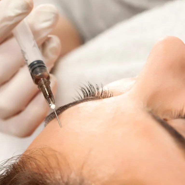 Close-up of a syringe near a person's forehead with eyes closed during a cosmetic procedure.