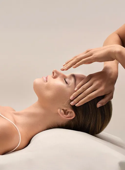 Woman lying down with eyes closed receiving a gentle forehead massage with hands.