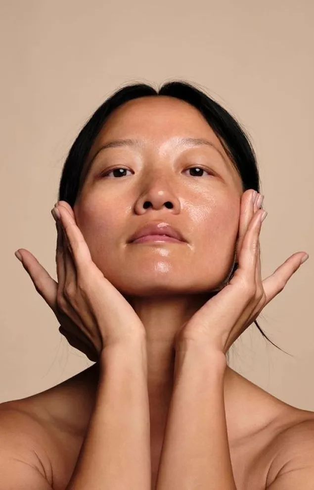 Woman with smooth skin gently cupping her face with both hands against a beige background.