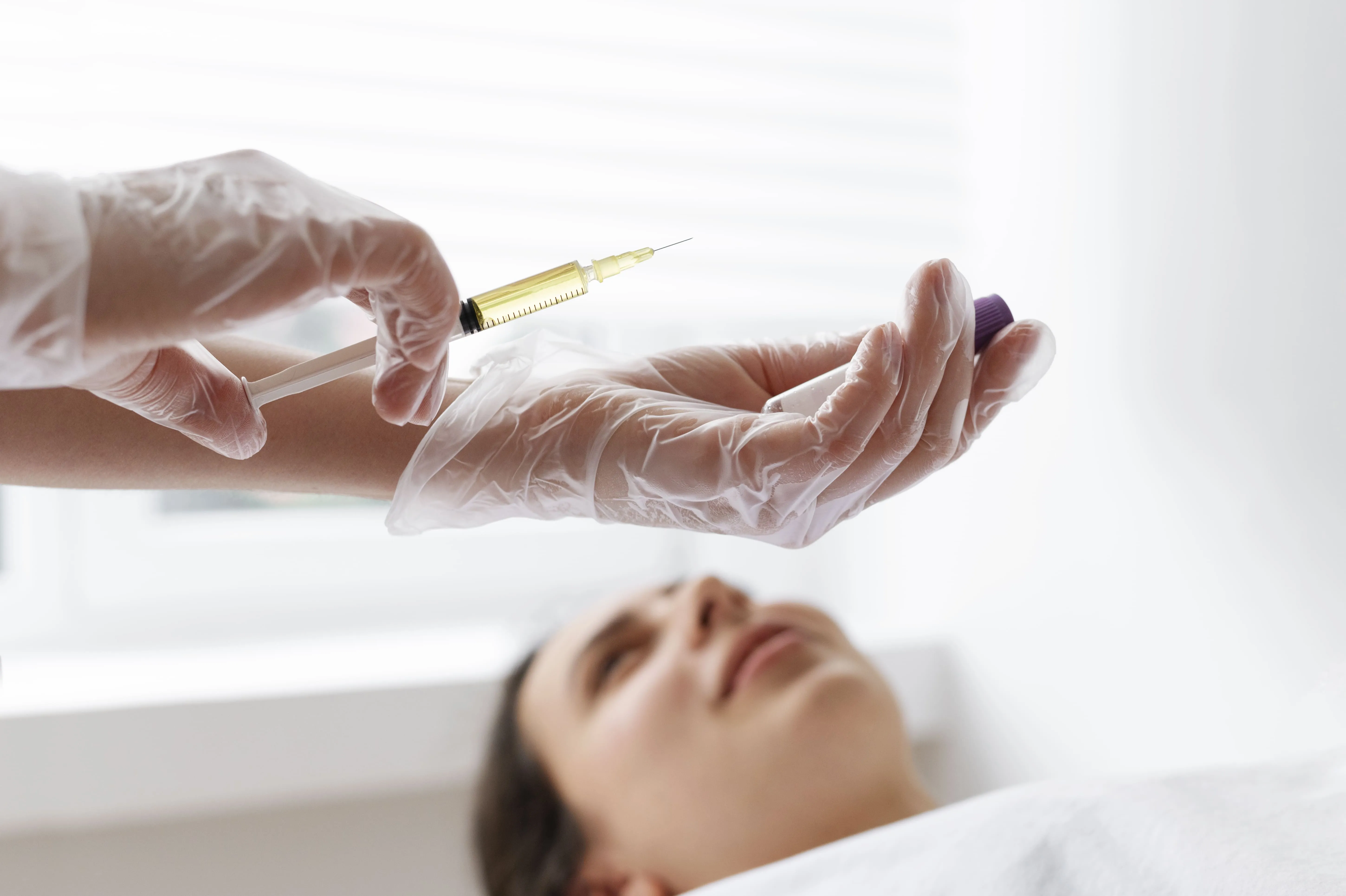 Gloved hands hold syringe and vial preparing injection with woman lying back, blurred in background.