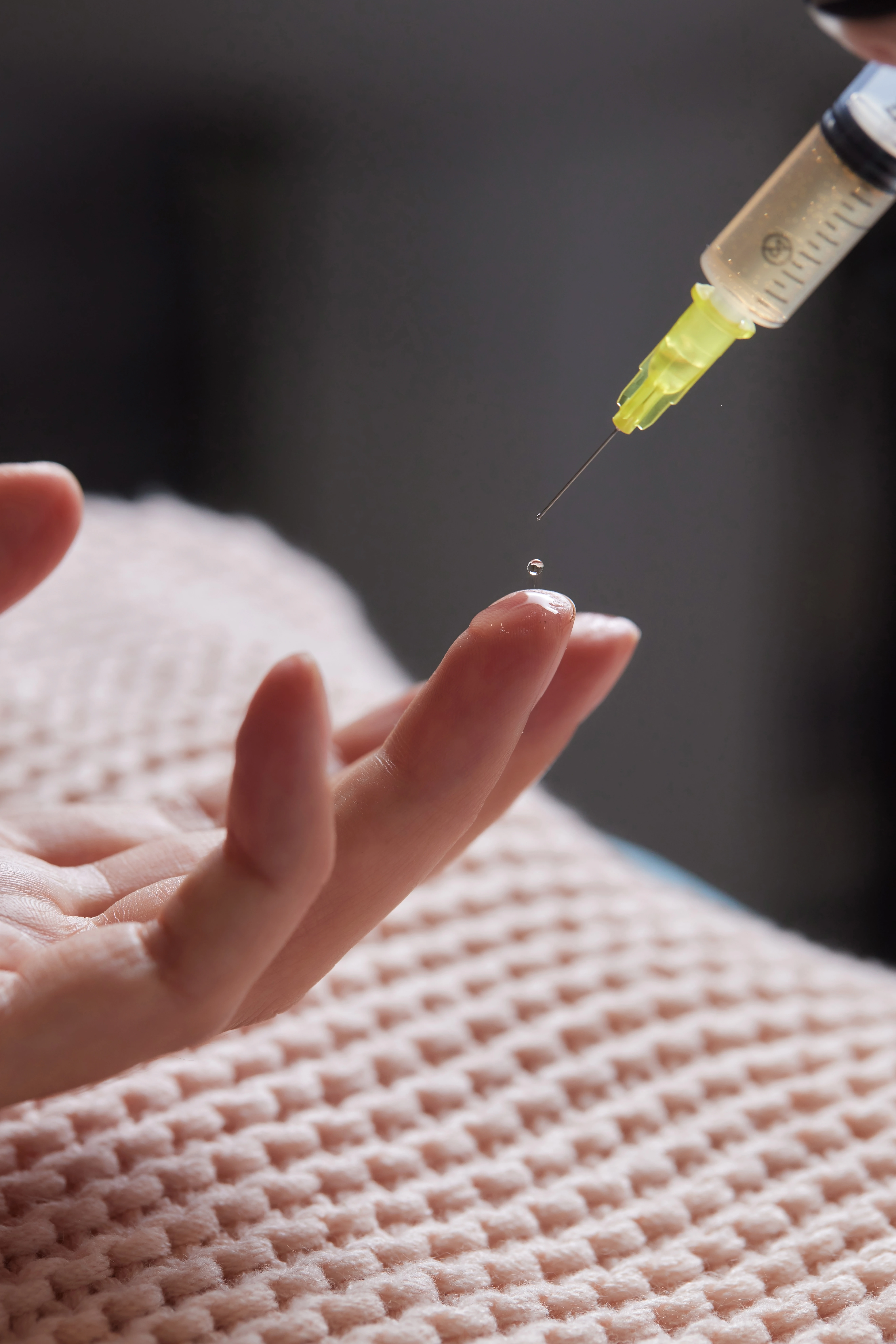 Droplet falling from syringe needle onto person's fingertip over a textured blanket.