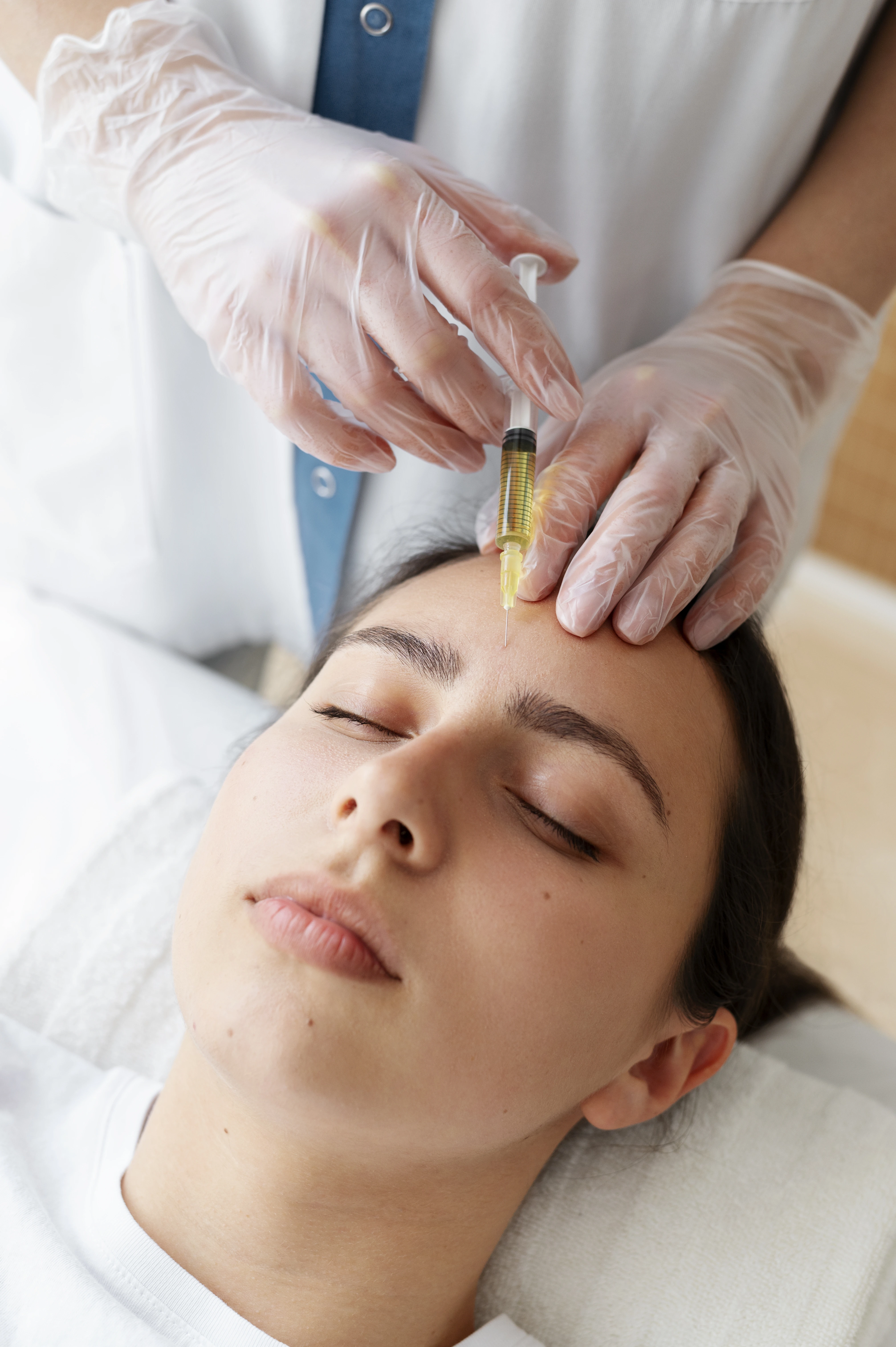 Gloved hands injecting a yellowish fluid into a woman's forehead during a cosmetic treatment.