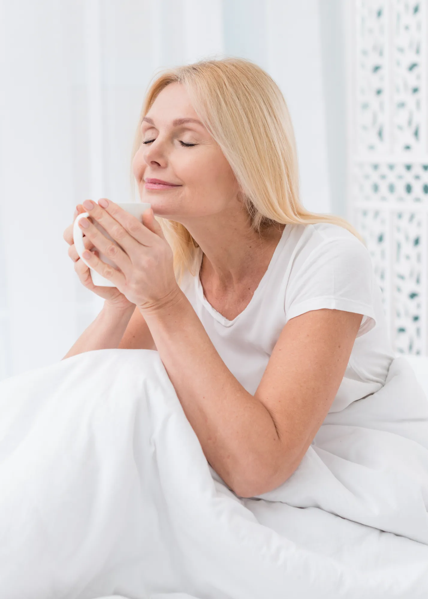 Older woman in white shirt enjoying the scent of a hot beverage while wrapped in a white blanket.