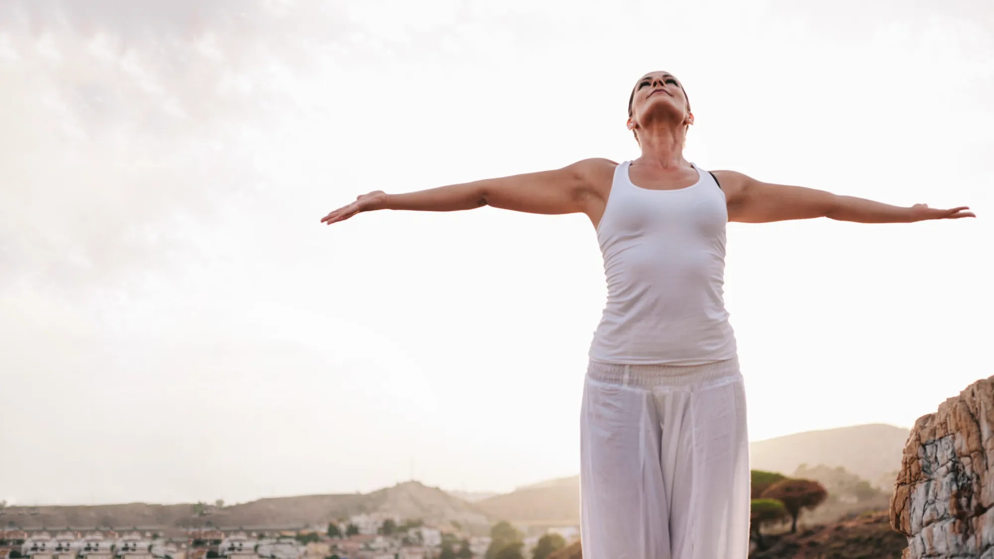 Woman in white clothes stretching arms wide outdoors with a town and hills in the background.