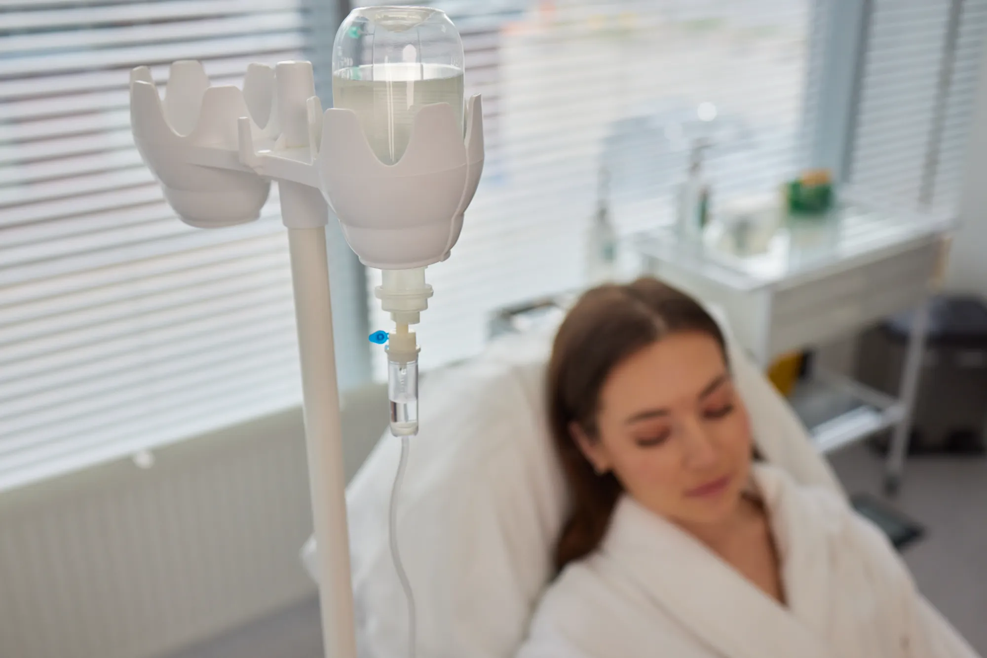 Close-up of an IV drip with a woman resting in a hospital bed in the background.