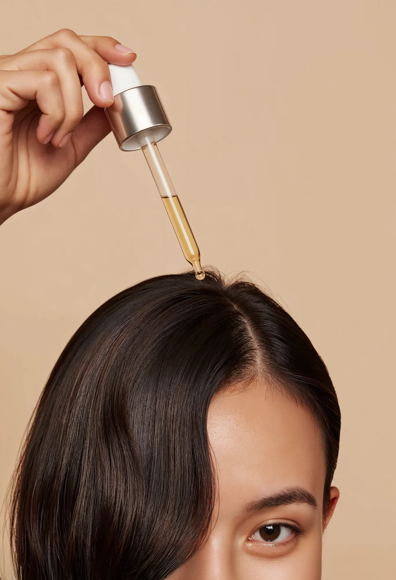 Close-up of hand applying hair serum with dropper to scalp of woman with dark hair.