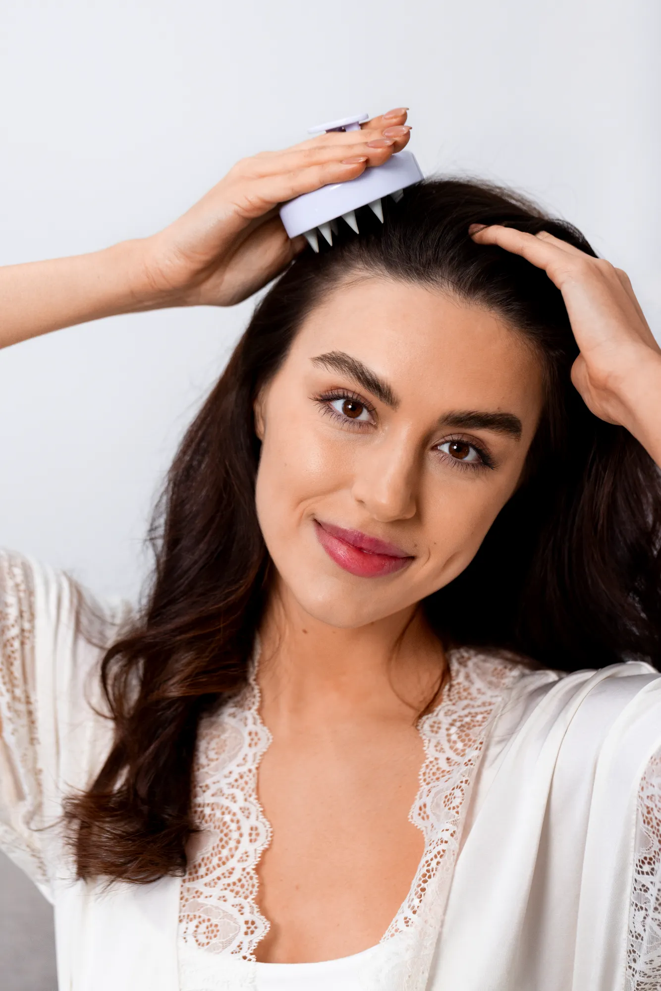 Smiling woman with dark hair massaging her scalp with a white scalp massager.