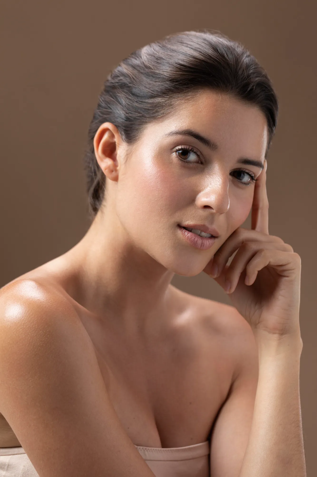 Close-up of a young woman with smooth skin posing against a brown background, resting her head on her hand.