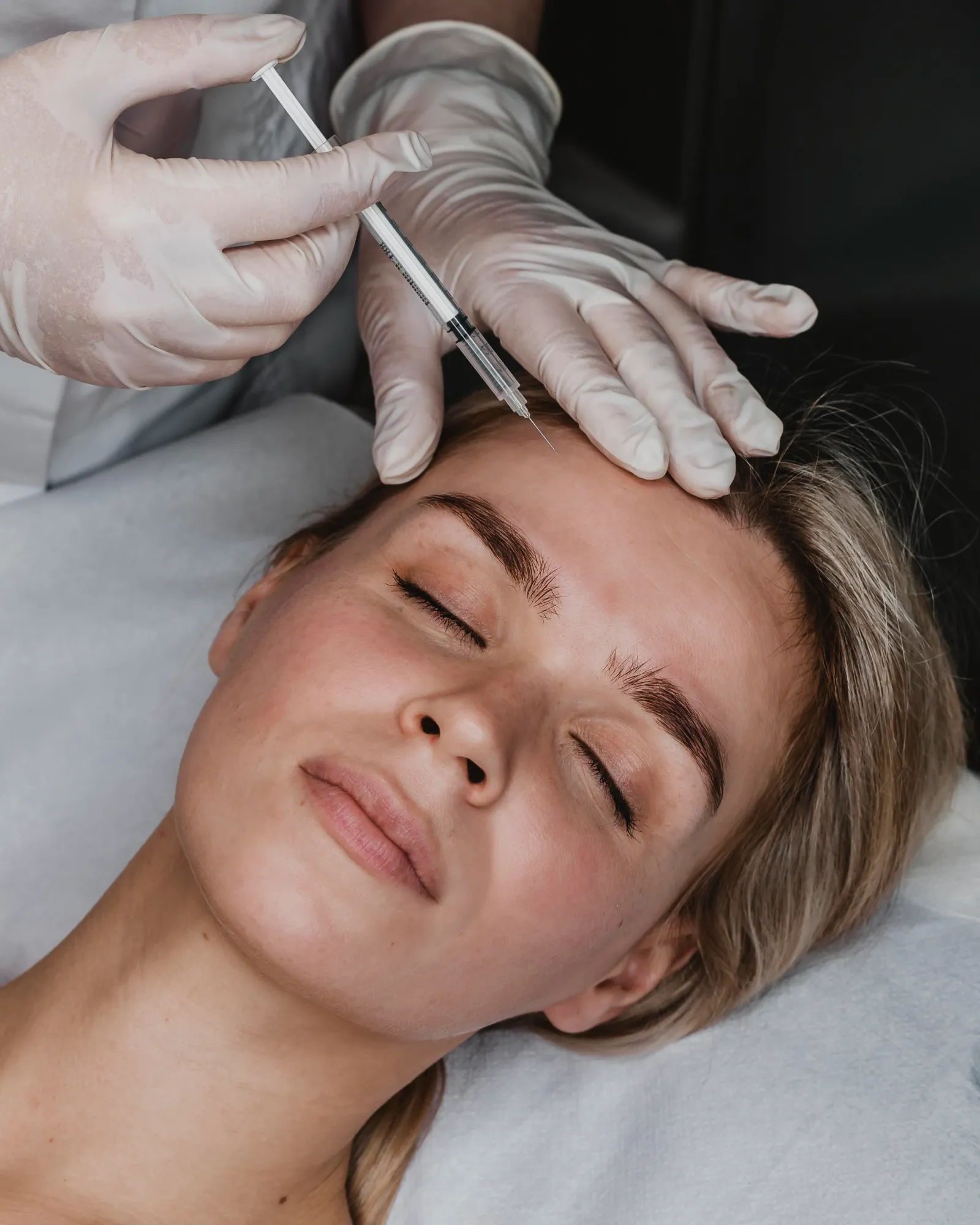 Gloved hands holding syringe near a woman's forehead preparing to give an injection.