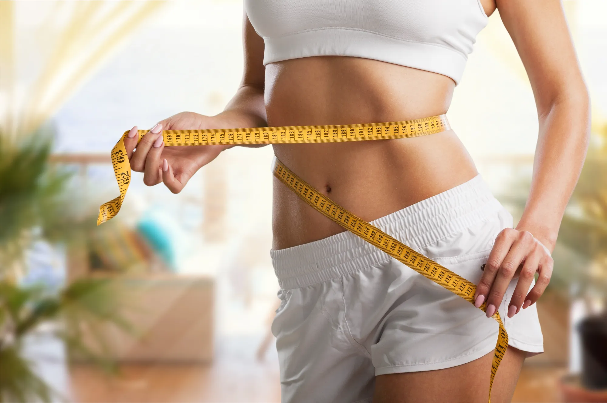 Woman in white sportswear measuring slim waist with yellow tape measure indoors.