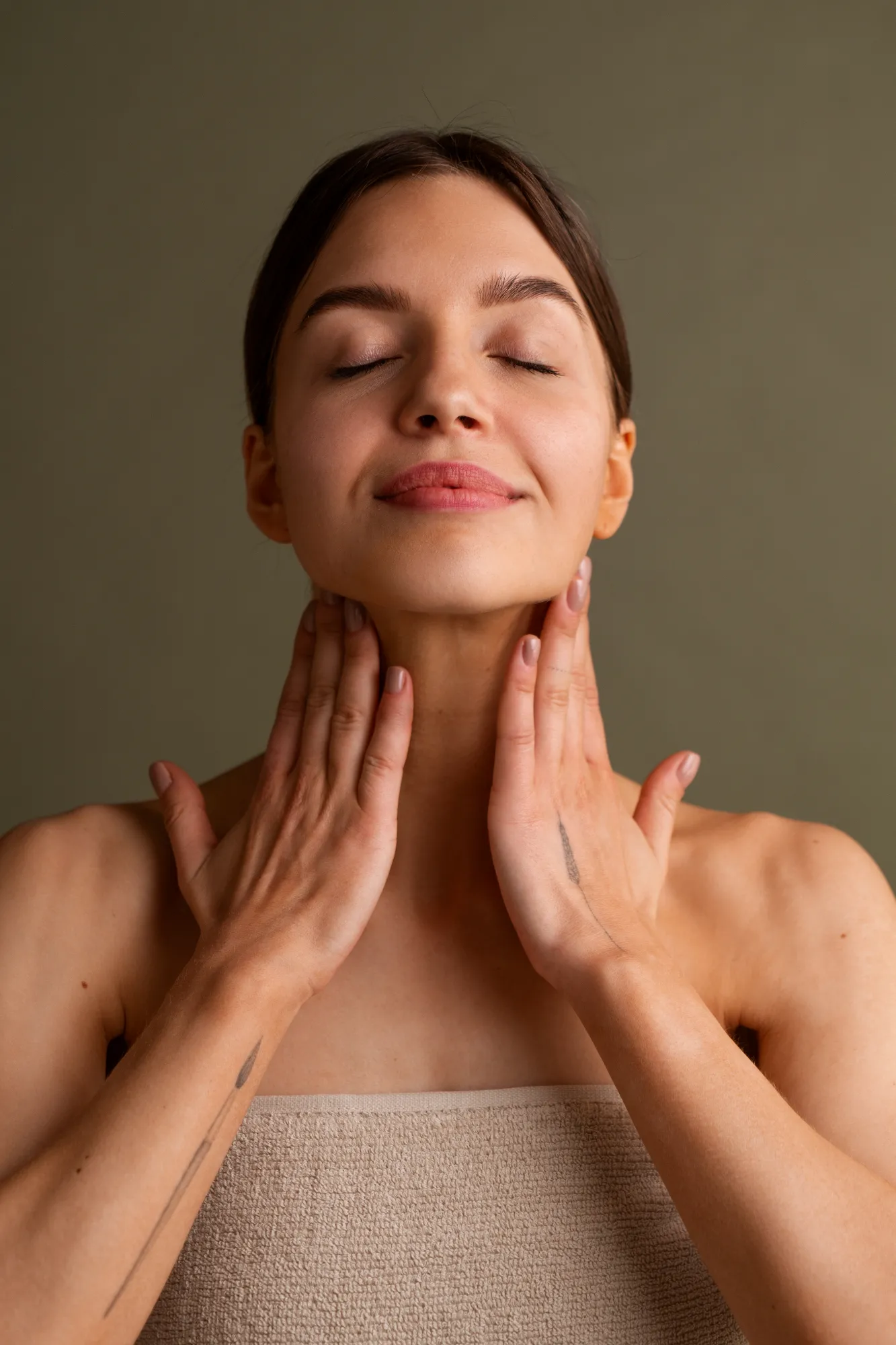 Woman with closed eyes massaging her neck with both hands, wearing a beige towel top.