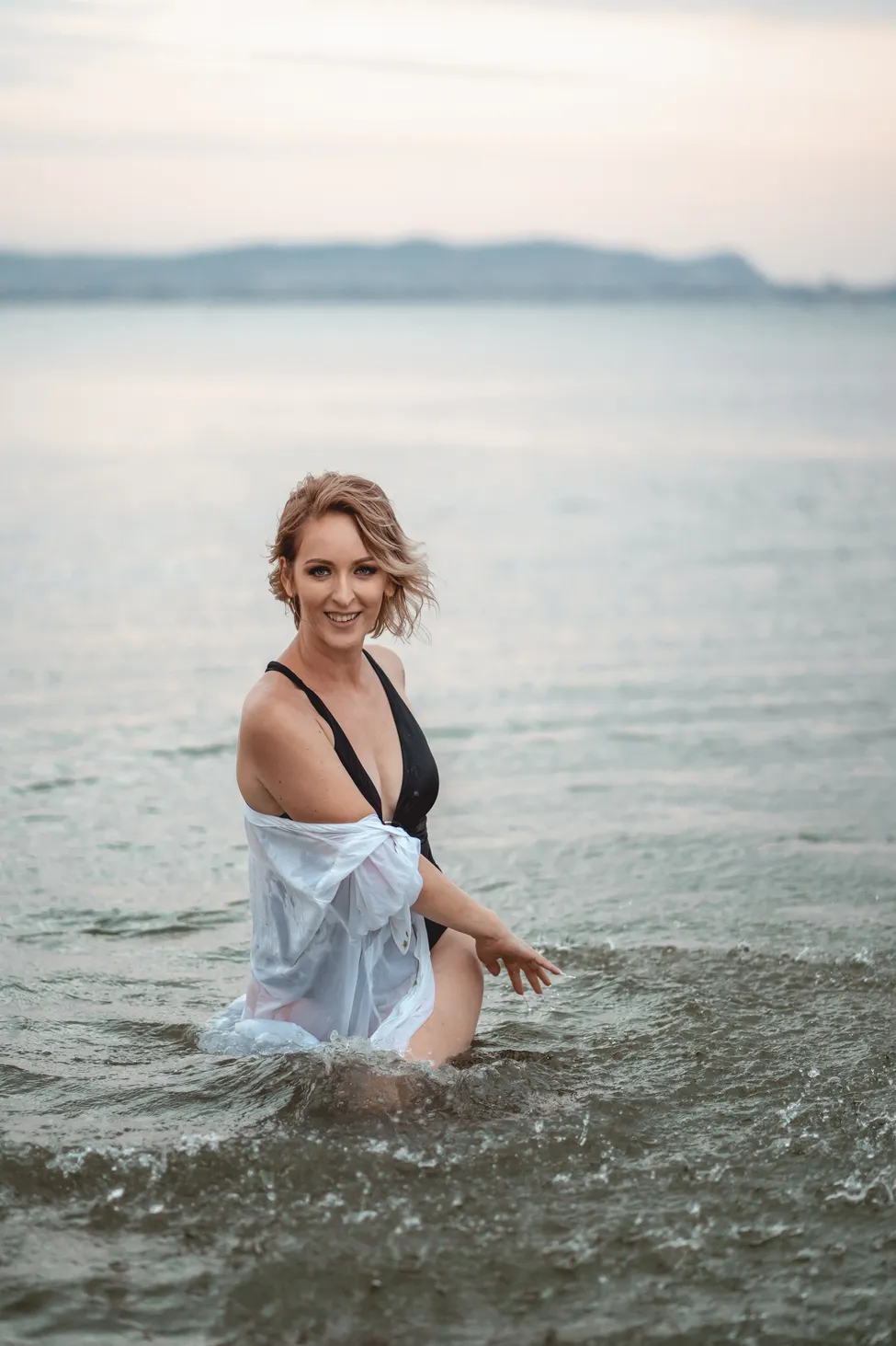 Smiling woman in black swimsuit and white shirt kneeling in shallow water at the beach.
