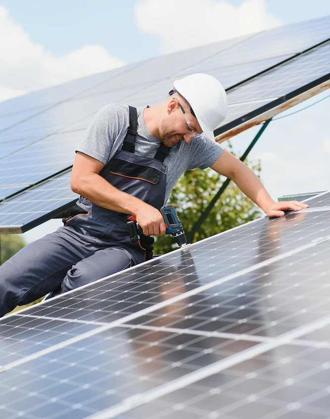 Dos trabajadores con cascos blancos y chalecos de seguridad instalando paneles solares en un techo mientras usan un ordenador portátil.