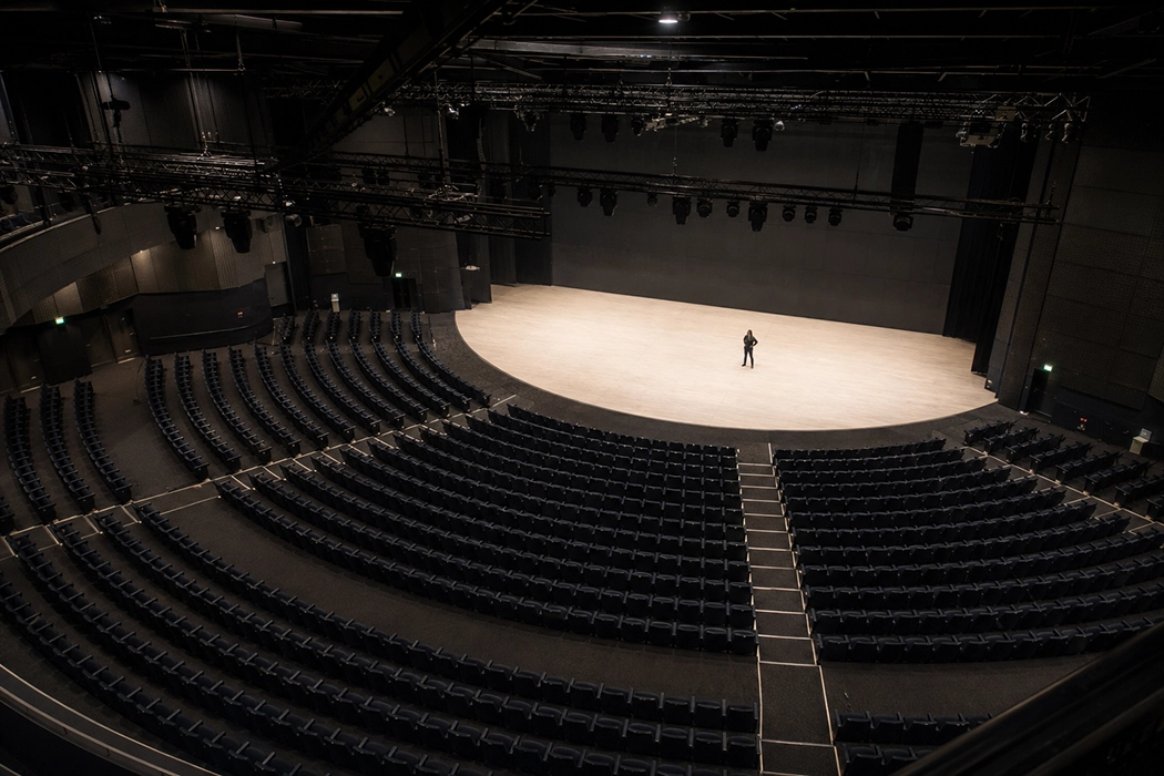 A lone person stands on a large empty stage in an auditorium with many empty seats.
