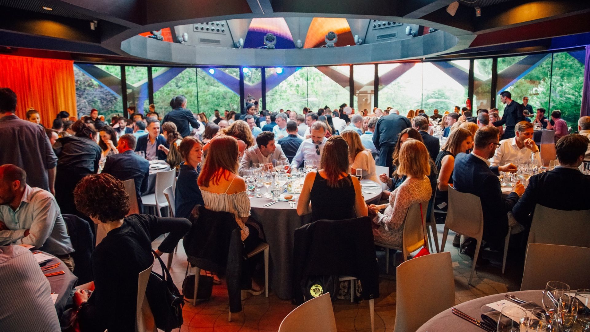 Large group of people sitting at multiple round tables in a modern indoor venue with large glass windows, engaging in conversation during a social or formal event.