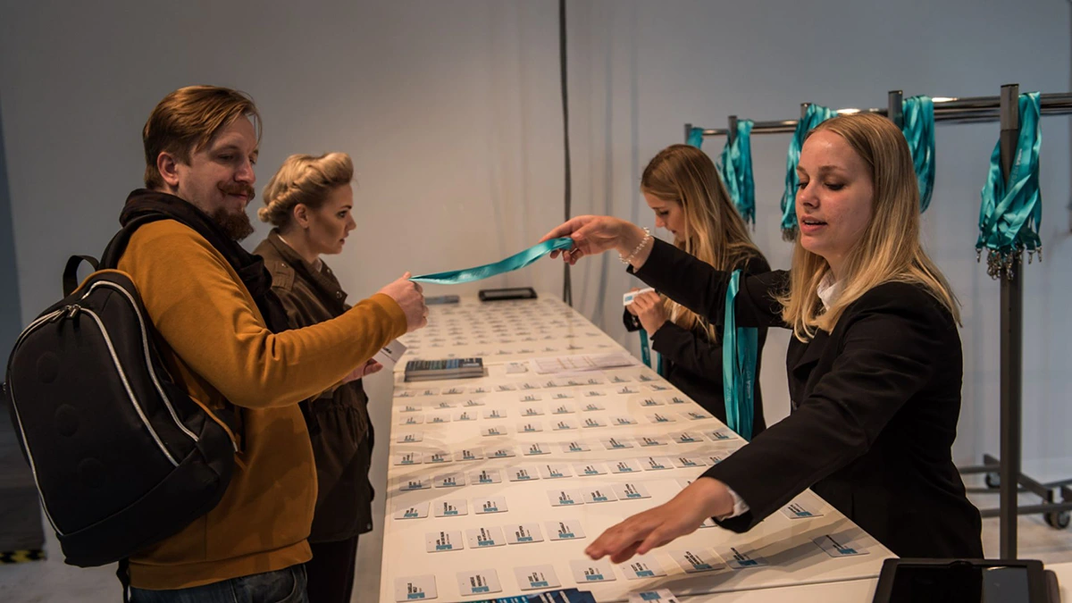 Two women behind a table handing out teal-colored lanyards to a man and a woman on the other side at an event registration or check-in area with many name badges laid out on the table.