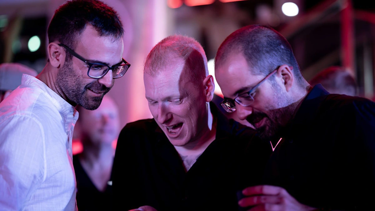 Three men looking down at something on a phone in a party setting.