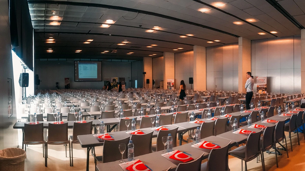 An empty conference venue, with rows of chairs facing a stage with a screen.