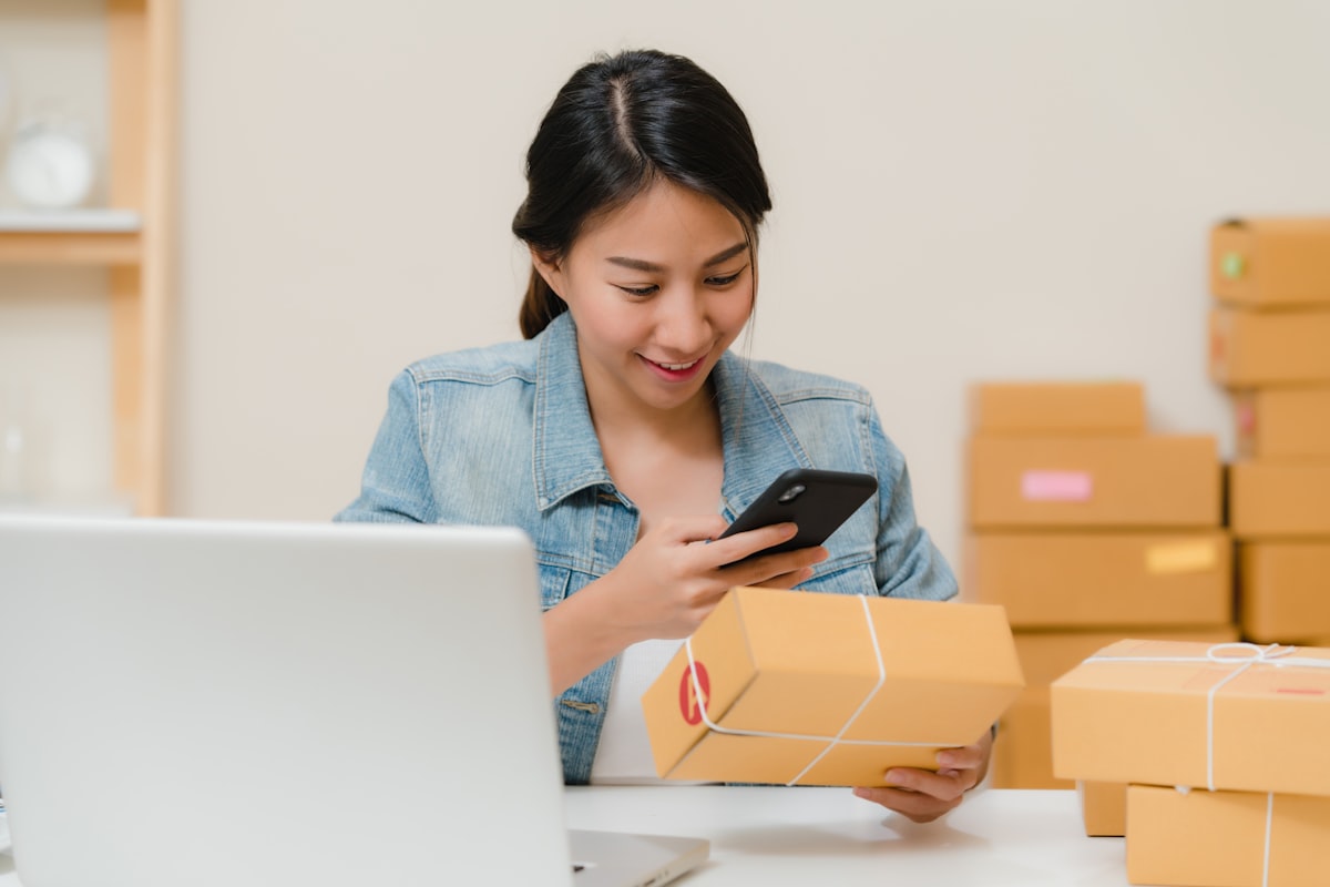 Woman checking package with phone near laptop and boxes. Photo by Rifki Kurniawan on Unsplash